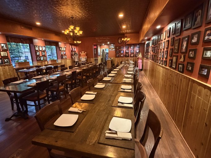 The interior of a restaurant shows long wooden communal tables set with white plates and napkins, wooden chairs, warm lighting from chandeliers, walls adorned with numerous framed black and white photographs, and a couple of figures standing in the background.