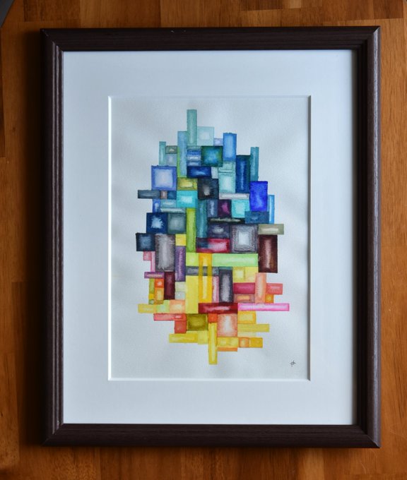 A framed abstract watercolor painting features an oval cluster of small colorful squares in blues, greens, yellows, reds, and pinks, mounted on a wooden surface.