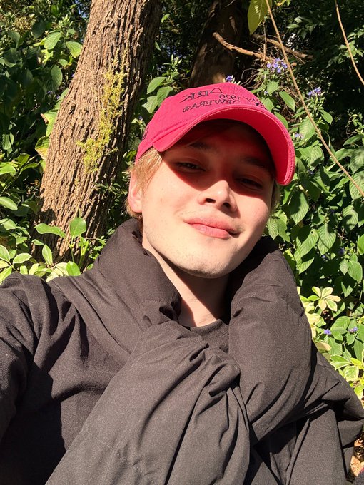A young man taking a selfie outdoors in front of green foliage, wearing a black hoodie and a pink baseball cap with the text 'VINYL DECO WEARS WU' visible on the front.