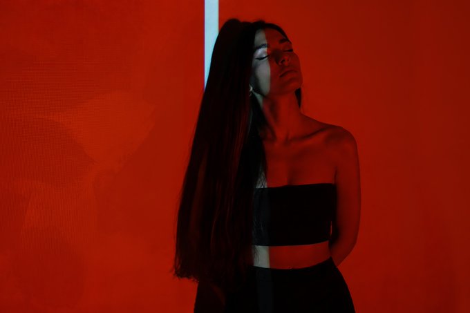 A woman with long dark hair wearing a black strapless top poses dramatically against a vivid red background illuminated by a vertical light strip.
