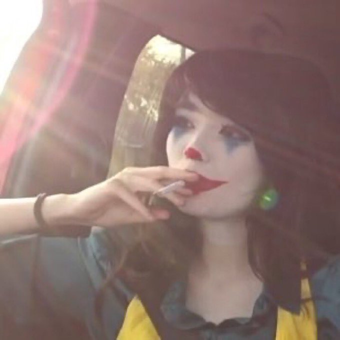A selfie of a person in clown-like makeup with a red nose and smeared lipstick, sitting in a vehicle with bright sunlight flaring in the background.