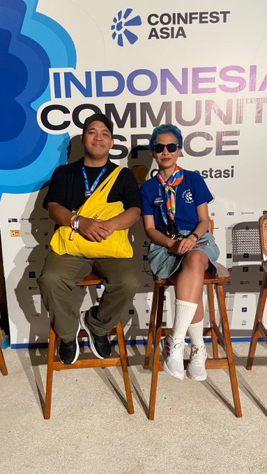 Two people sit on stools in front of a 'Coinfest Asia Indonesia Community Space' banner; the man wears a black shirt, olive pants, and a yellow crossbody bag, while the woman has blue hair, sunglasses, a blue shirt, and white knee-high socks.