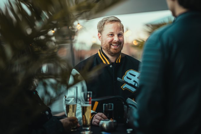 A bearded man smiles while wearing a black and yellow varsity-style jacket featuring a prominent "SF" logo, identified as Solflare merchandise from a web3 gathering with drinks on the table. The image captures the casual, social atmosphere referenced in the post's #GMerchMonday feature.