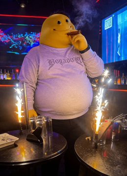 A person in a large yellow smiley face mascot costume wearing a sweatshirt reading 'Boystown' appears to be smoking, with sparklers burning on a bar counter in the foreground.