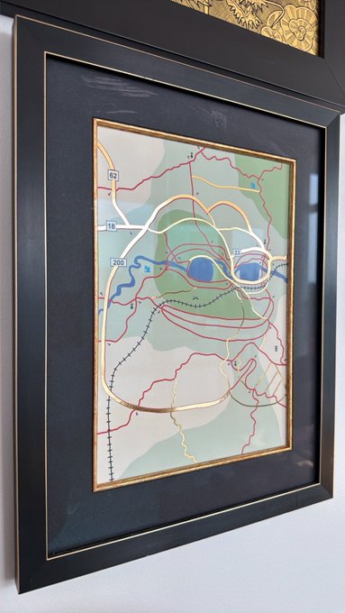 A framed piece of artwork featuring an abstract face design overlaid on what appears to be a road or map, with visible route numbers like '62', '10', '200', and '11', displayed in a black and gold frame.