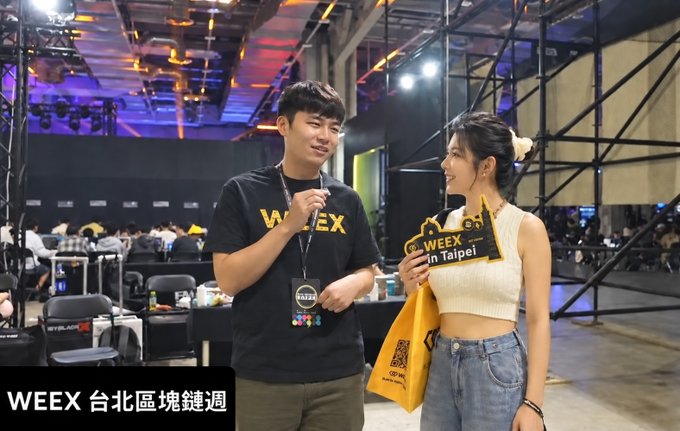 A man wearing a black 'WEEX' t-shirt interviews a smiling woman in a white crop top at what appears to be a crypto event, with text reading 'WEEX in Taipei' on her sign and 'WEEY 台北區塊鏈週' at the bottom.