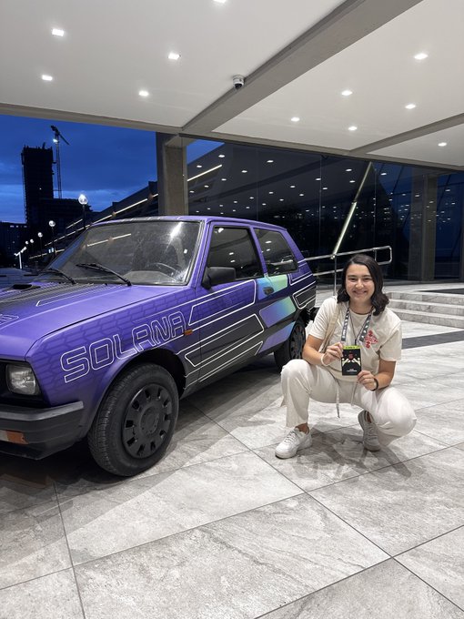 A smiling person crouches next to a purple hatchback car with 'SOLANA' written on its side, in what appears to be an indoor event space.