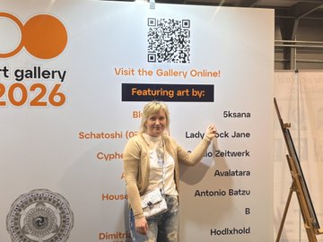 The image features a woman standing next to a white wall with an orange sign that reads "Visit the Bitcoin Art Gallery Online @BTCPrague". The woman appears to be posing for a photo, possibly at an art exhibition or event related to Bitcoin art.