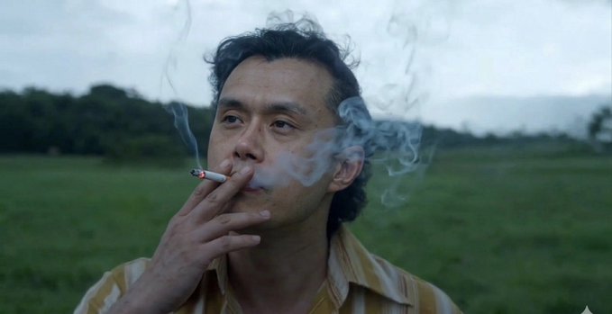 A man in a yellow and white striped shirt smokes a cigarette outdoors, exhaling smoke against a backdrop of a green field and overcast sky.