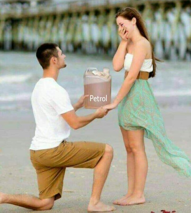 The image shows a man kneeling down next to a woman, presenting her with a can of Petrol as part of their engagement moment. They are both barefoot, enjoying each other's company at the beach. The couple appears to be celebrating their love and commitment in this unique and romantic setting.
