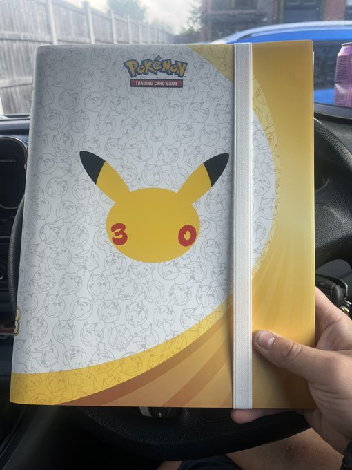 A hand holds a Pokémon Trading Card Game 30th anniversary Pikachu binder inside a car, featuring yellow and white coloring with Pikachu's face and the number '30' on the cover.