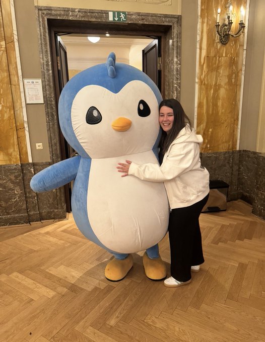 A woman poses warmly with a large blue and white Pudgy Penguin mascot in an elegant marble-walled venue, capturing a celebratory moment tied to the Schleich® Collectible Figurines Series 1 debut at Spielwarenmesse in Nuremberg. The plush penguin, a centerpiece of the event, embodies the brand's playful European launch amid the toy fair's 2,300+ exhibitors and nearly 59,000 trade visitors.