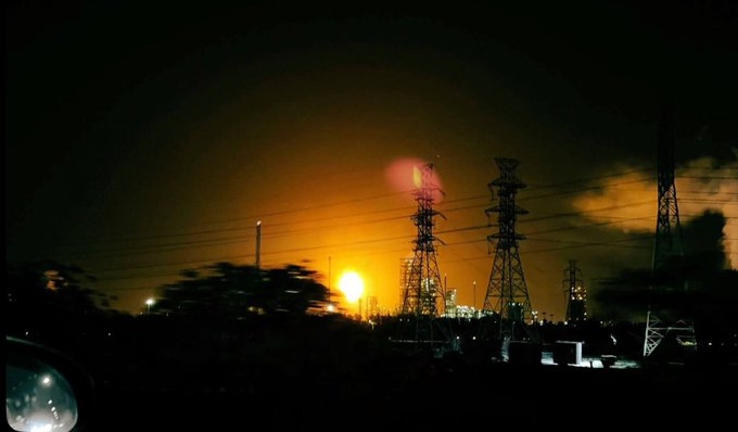 A nighttime photograph of an industrial facility with tall power transmission towers silhouetted against an orange glow from what appears to be a fire or flare in the background.