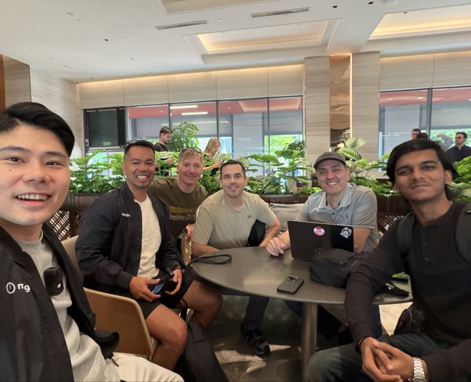 Six people pose for a group photo around a table in what appears to be a hotel or conference lounge, with a man on the left taking a selfie. The setting, with its modern interior, plants, and business-casual attire, reflects the Token 2049 conference gathering mentioned in the caption, as the group seems to have arrived in Singapore for the event.
