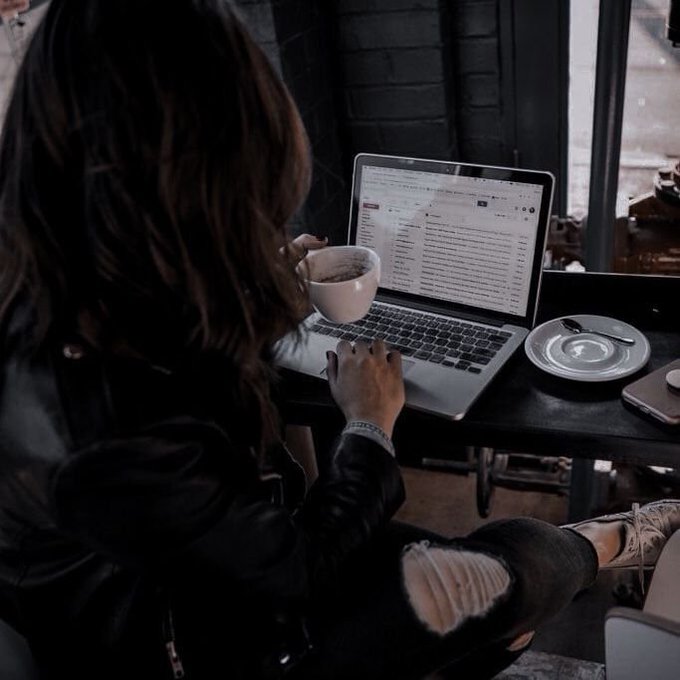 A person with long dark hair sits at a cafe table working on a MacBook laptop while holding a white coffee mug, with a saucer and spoon visible on the dark wooden table.