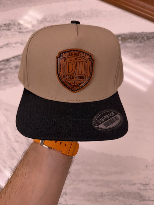 A hand wearing an orange watch holds up a tan and black snapback hat featuring a brown leather patch that reads '589 DAYS OF DH DEGEN HOURS EST. 2024' and a 'SNAPBACK' sticker on the brim.
