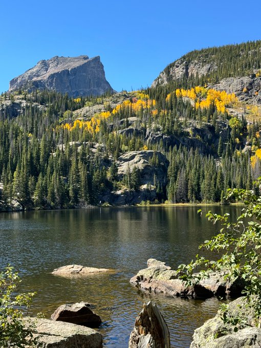 A landscape photograph of a mountain lake surrounded by rocky cliffs and evergreen trees, with patches of yellow autumn foliage visible on the slopes.