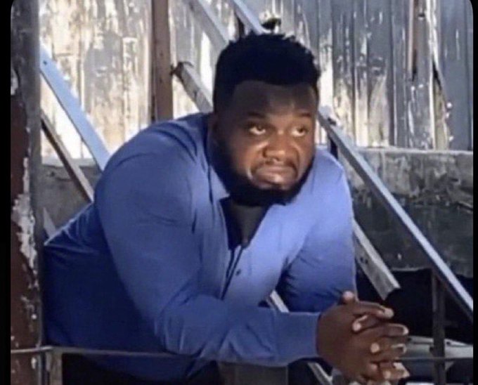 A man in a blue long-sleeve shirt leaning on a railing with his hands clasped, looking forward with a serious expression in what appears to be a rustic or industrial setting.