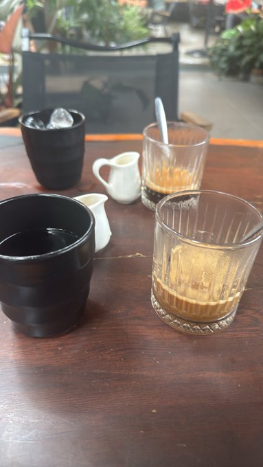 Two cups of Vietnamese coffee (cà phê sữa đá) with small cream pitchers sit on a wooden table at an outdoor café, where a man is enjoying his morning coffee with his wife as referenced in the post's greeting.