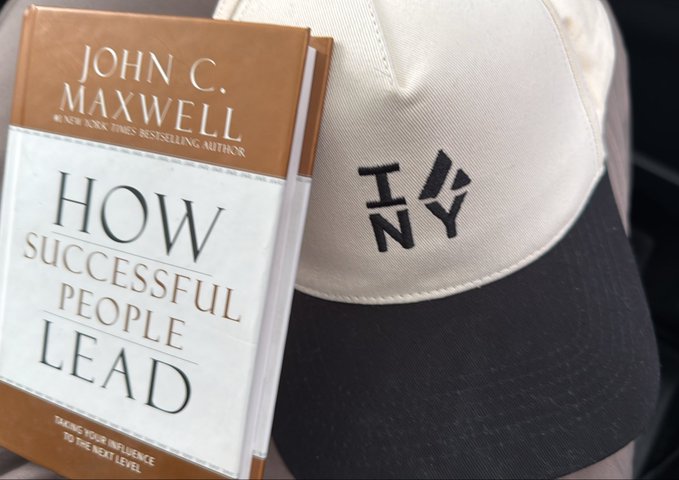 A copy of John C. Maxwell's book 'How Successful People Lead: Taking Your Influence to the Next Level' rests against a cream and black baseball cap with an embroidered logo.