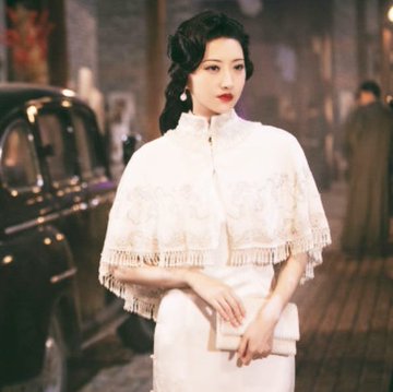 A woman in an elegant white vintage qipao dress with a lace and fringe shawl stands holding a clutch purse on a cobblestone street at night next to a vintage car.
