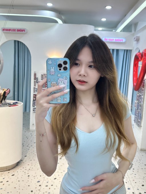 A young woman with long dark hair takes a mirror selfie in a store using a phone with a blue bunny-decorated case, wearing a light blue top.