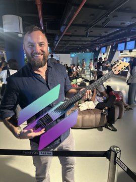 A bearded man smiling while holding a purple and green multi-colored electric guitar at what appears to be a gaming event.