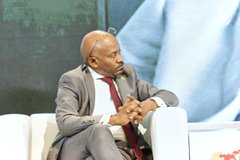 A photo of Mike Tyson in a gray suit and red tie sitting thoughtfully on a white chair at what appears to be a panel or conference event.