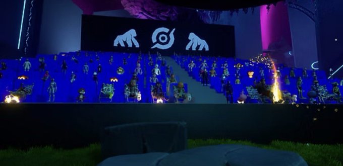 A virtual stage in a digital environment filled with blue seats and various avatar characters, featuring a large screen displaying two hand logos and an eye symbol.