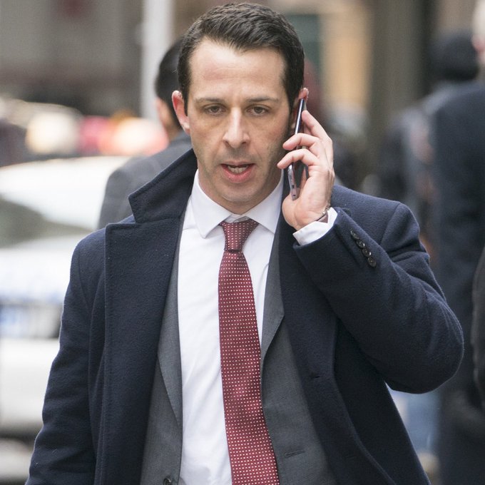 A man in a navy overcoat and burgundy tie walks down a city street, talking on his phone with a serious expression while an out-of-focus crowd and car are visible behind him—capturing the frantic, distracted energy of someone canceling plans over an absurdly niche tech obsession.