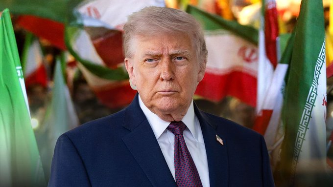 The image features a man dressed in a suit standing in front of a group of flags. The man appears to be wearing a tie with purple and white colors. The flags behind him are green, red, and white, creating an interesting visual contrast against each other. The scene seems to be set in a room or a public space where the flags are displayed prominently.