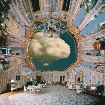 The image features an indoor room with a large chandelier hanging from the ceiling, creating a beautiful ambiance. The room has a high ceiling adorned with ornate decorations that give it an elegant appearance. There are several chairs placed around the room, some of which are occupied by people who seem to be enjoying themselves. A dining table is also present in the room, suggesting that this space could serve as both a social gathering spot and a place for meals. The image appears to have been taken during an auction or event where multiple pieces were up for bidding.