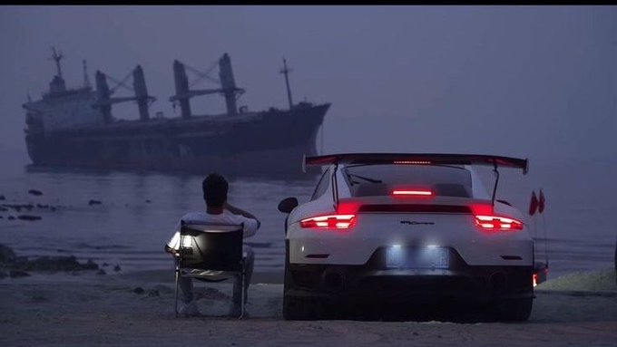A man sits in a folding chair next to a white Porsche sports car on a beach at dusk, with a large cargo ship anchored in the misty water behind them.
