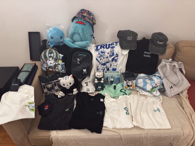 A collection of various merchandise items, including plush toys, t-shirts, hats, backpacks, and bags, arranged on a beige couch.