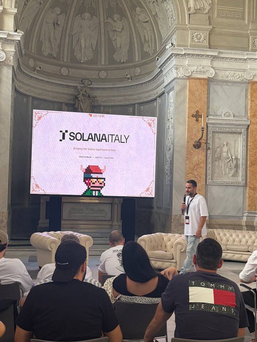 The image shows a man standing at a podium, giving a speech or presentation about the future of @solana Italy. The audience consists of several people sitting in chairs around the room, attentively listening to the speaker. A large screen displaying the @solana logo is visible behind the speaker, emphasizing the theme of the event.