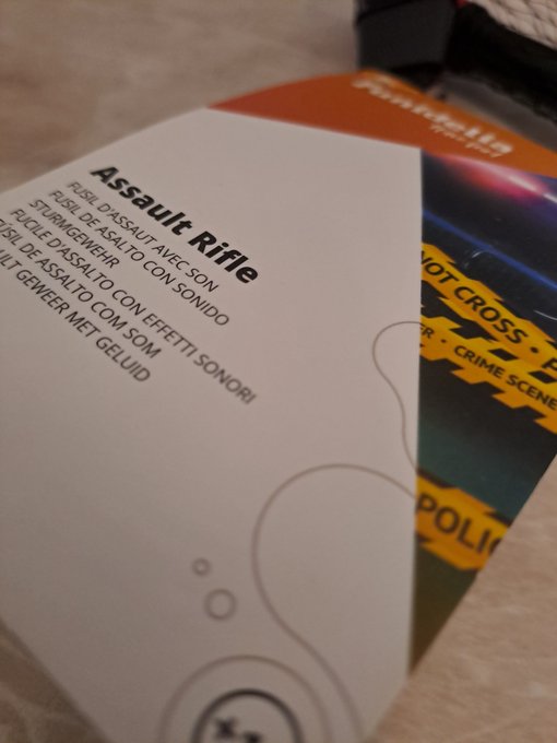 A close-up of a card with the title "Assault Rifle" and translations in multiple languages ("FUSIL D'ASSAUT AVEC SON", "STURMGEWEHR"), layered over a "DO NOT CROSS" crime scene themed card.