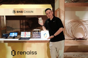 A man stands in a booth labeled 'BNB CHAIN' and 'renaiss' displaying several laptops and holding a product box.