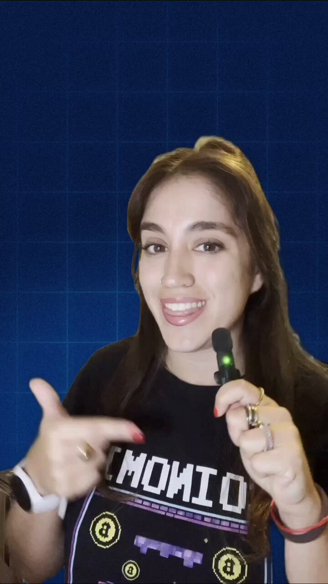 The image shows a woman wearing a black t-shirt with white text, smiling at the camera and pointing her finger towards it while holding a device that appears to be an e-cigarette, possibly an electronic cigarette or vaporizer. The photo is set against a blue background, which emphasizes the subject of the image. There are no other visible objects in the picture, and the woman seems to be engaging with the viewer through her body language and facial expression. It's unclear what specific topic or context the woman might be discussing in relation to this image, but it could possibly be related to a post about electronic cigarettes, e-cigarettes, or vaping.