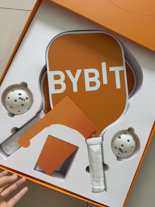 An overhead view of an opened orange and white Bybit branded gift box containing a white ping pong paddle, two balls, and a white grip handle accessory.