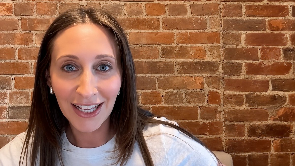 A woman with long brown hair wearing a white shirt smiles at the camera against a brick wall background.