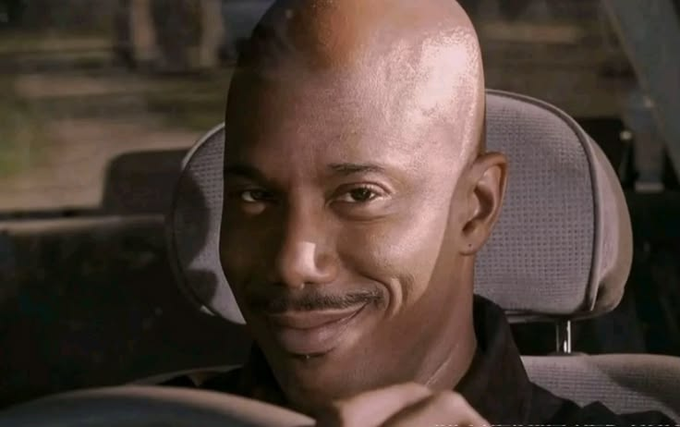 A man with a shaved head and small mustache smiles subtly while sitting in what appears to be a car seat.