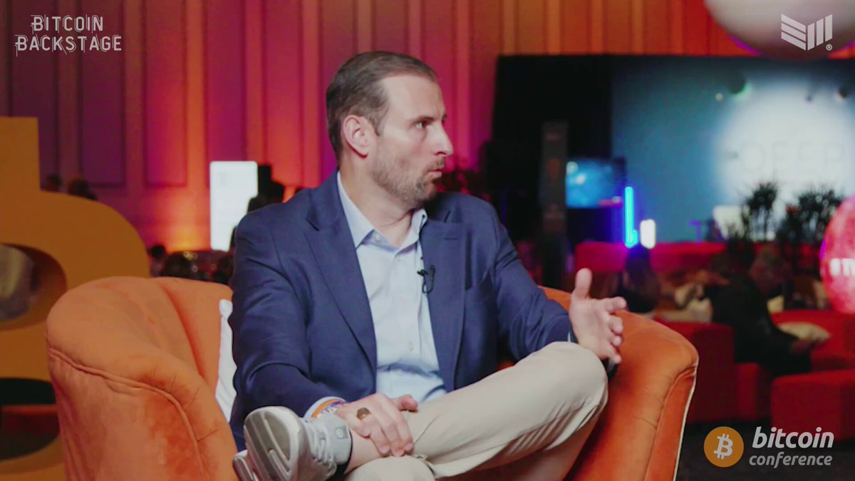 The image shows a man sitting down, wearing a blue suit jacket and khaki pants. He appears to be at a Bitcoin conference or event, as indicated by the text "White House BitCoin CHIEF PATRICK WITT BACKSTAGE @THEBITCOIN CONFERENCE". The man is seated in an orange chair with his legs crossed, giving him a relaxed yet professional appearance.