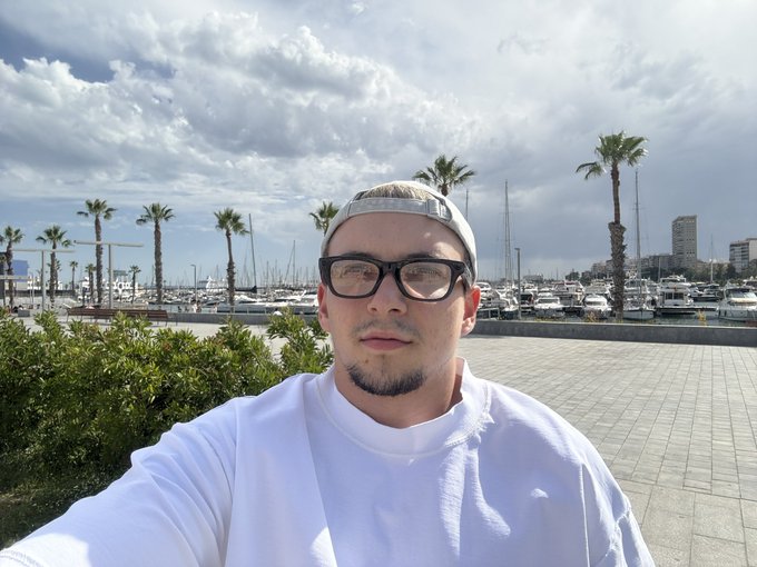 A man with glasses, a beard, and a backwards cap takes a selfie at a marina with palm trees, sailboats, and a cloudy sky in the background.