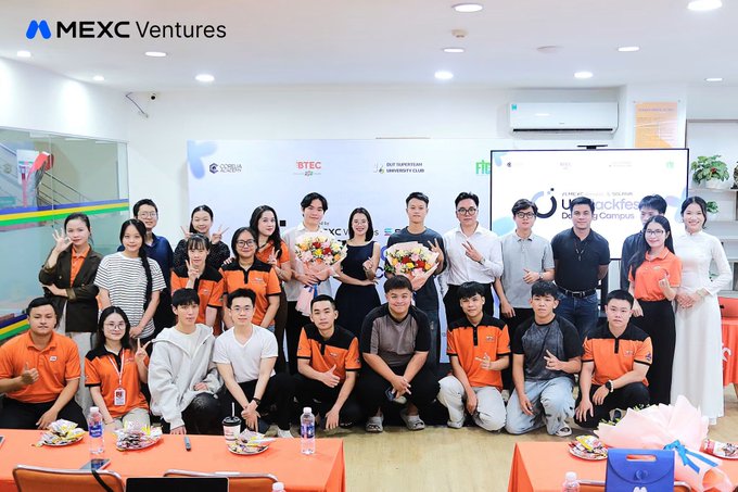 A group photo of approximately 25 people at a MEXC Ventures event, with attendees wearing a mix of orange shirts and formal attire, posing in front of banners including 'MEXC Ventures', 'BTEC', and 'FPT University' logos.