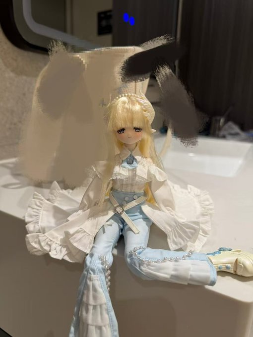 A ball-jointed doll (BJD) with blonde hair and pointed ears sits on a bathroom counter, dressed in an intricate white and light blue outfit with frills, lace, and beaded accents.