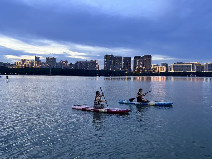 Two people paddleboarding on calm water at dusk, with a city skyline of illuminated high-rise buildings in the background.