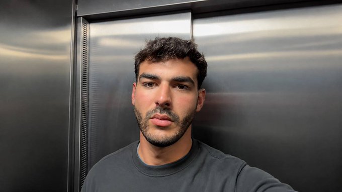 A young man with curly dark hair and a short beard poses for a close-up selfie inside an elevator with metallic walls, wearing a dark gray t-shirt. The casual, direct gaze at the camera fits the "Good Morning" energy of the post asking followers to share their $SOL crypto wallet addresses.