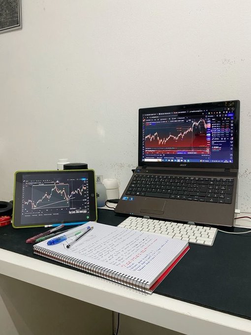 A desk setup features an Acer laptop and a tablet, both displaying trading candlestick charts, alongside a spiral notebook with handwritten notes, pens, and a white keyboard.
