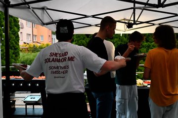 A vendor wearing a 'She Said No I Said Solanor Me Sometimes I Wish Her' t-shirt serving food at an outdoor market stall under a white tent canopy.