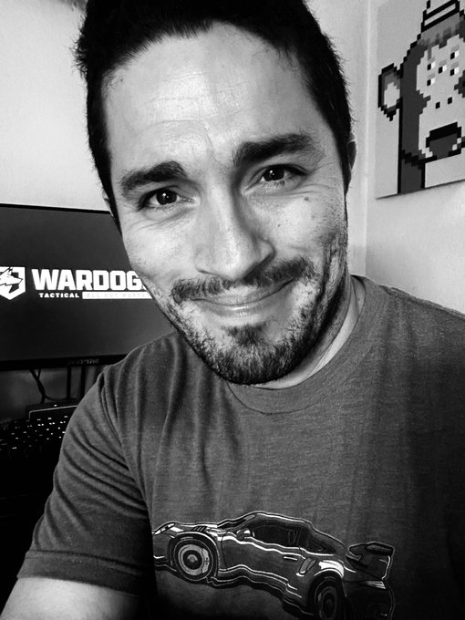 A black-and-white selfie shows a smiling man seated near a monitor displaying the “WARDOGG TACTICAL” logo and name; he wears a T-shirt with a sports car graphic.